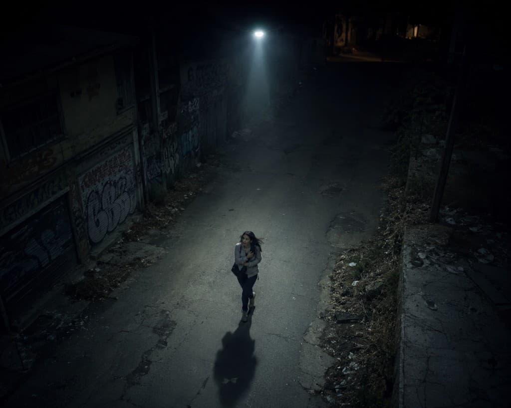 A woman walking through a dark alley at night, illuminated by a bright overhead spotlight